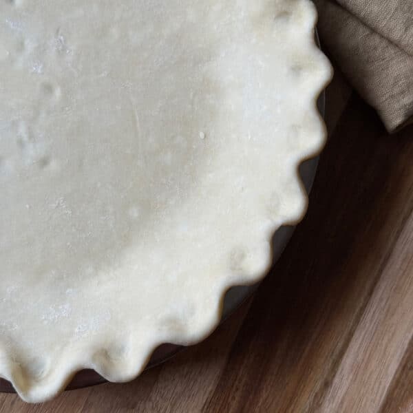 Unbaked pie with crimped edges in a pie dish on a wooden surface, partially covered by a brown cloth.