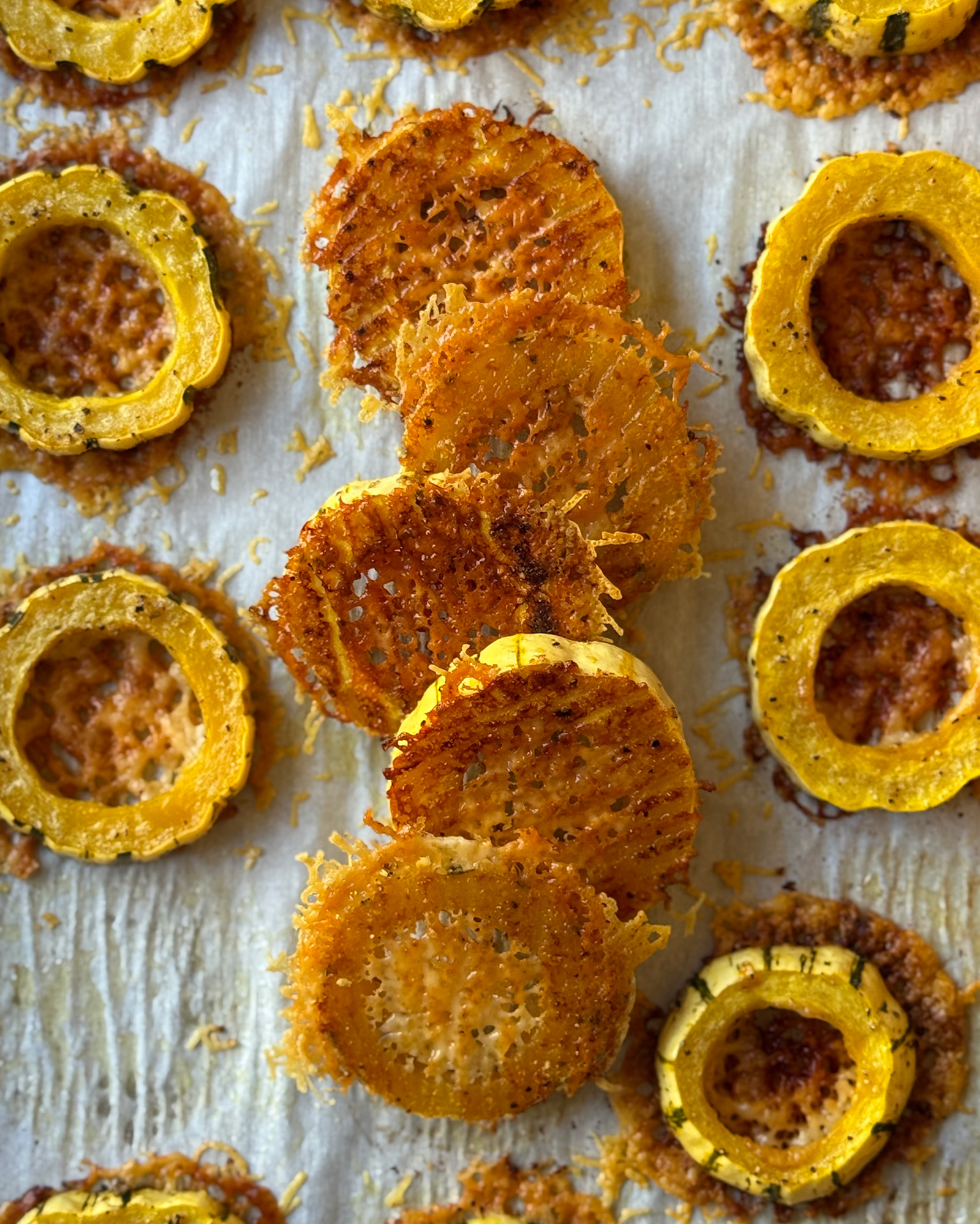 Baked delicata squash rings flipped and overlapping down the center of a parchment-lined baking sheet, showing their crispy Parmesan crusts, with upright rings on either side.