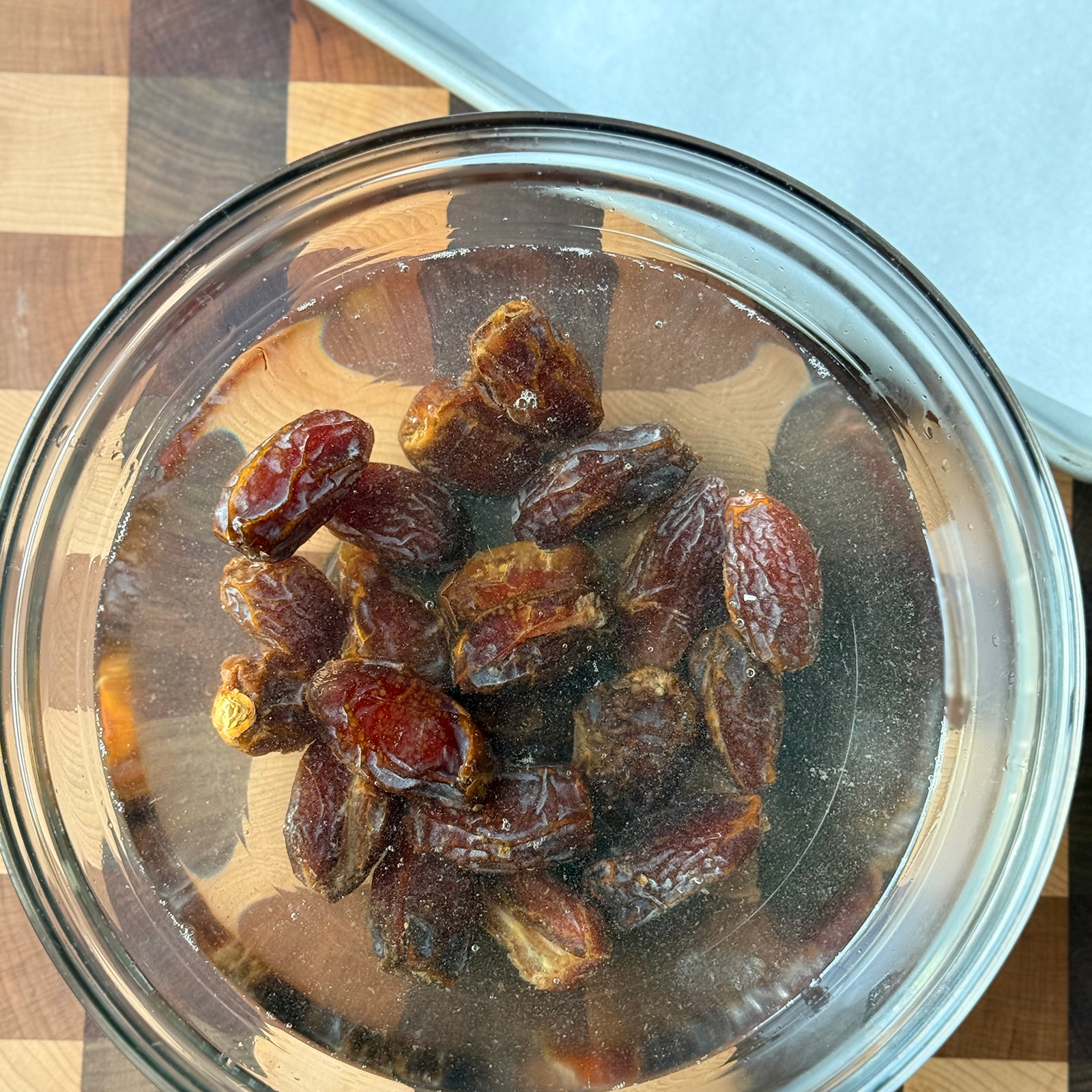Medjool dates soaking in warm water in a glass bowl on a checkered cutting board.
