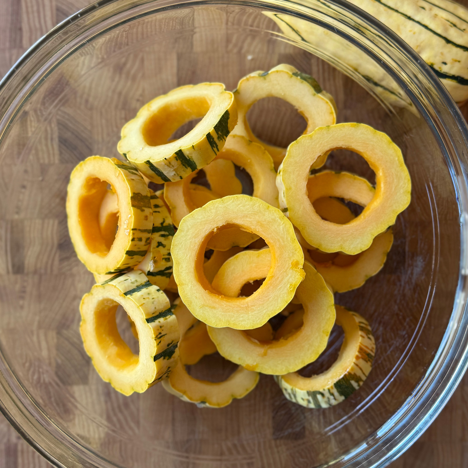 Delicata squash sliced into rings with the seeds scooped out, in a glass bowl beside whole delicata squash.