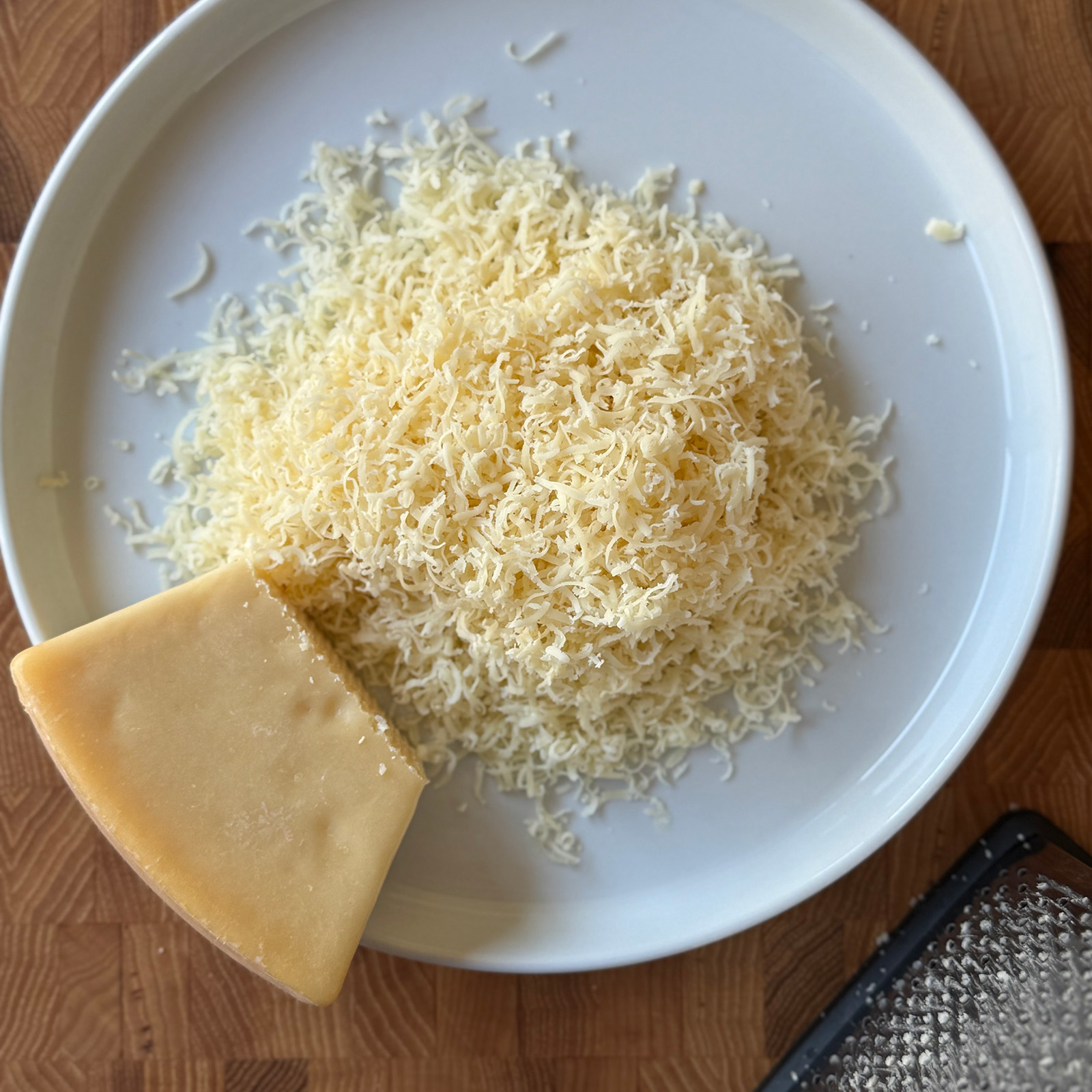 Freshly grated vegetarian Parmesan on a white plate with a wedge of cheese and a box grater on a wooden cutting board.