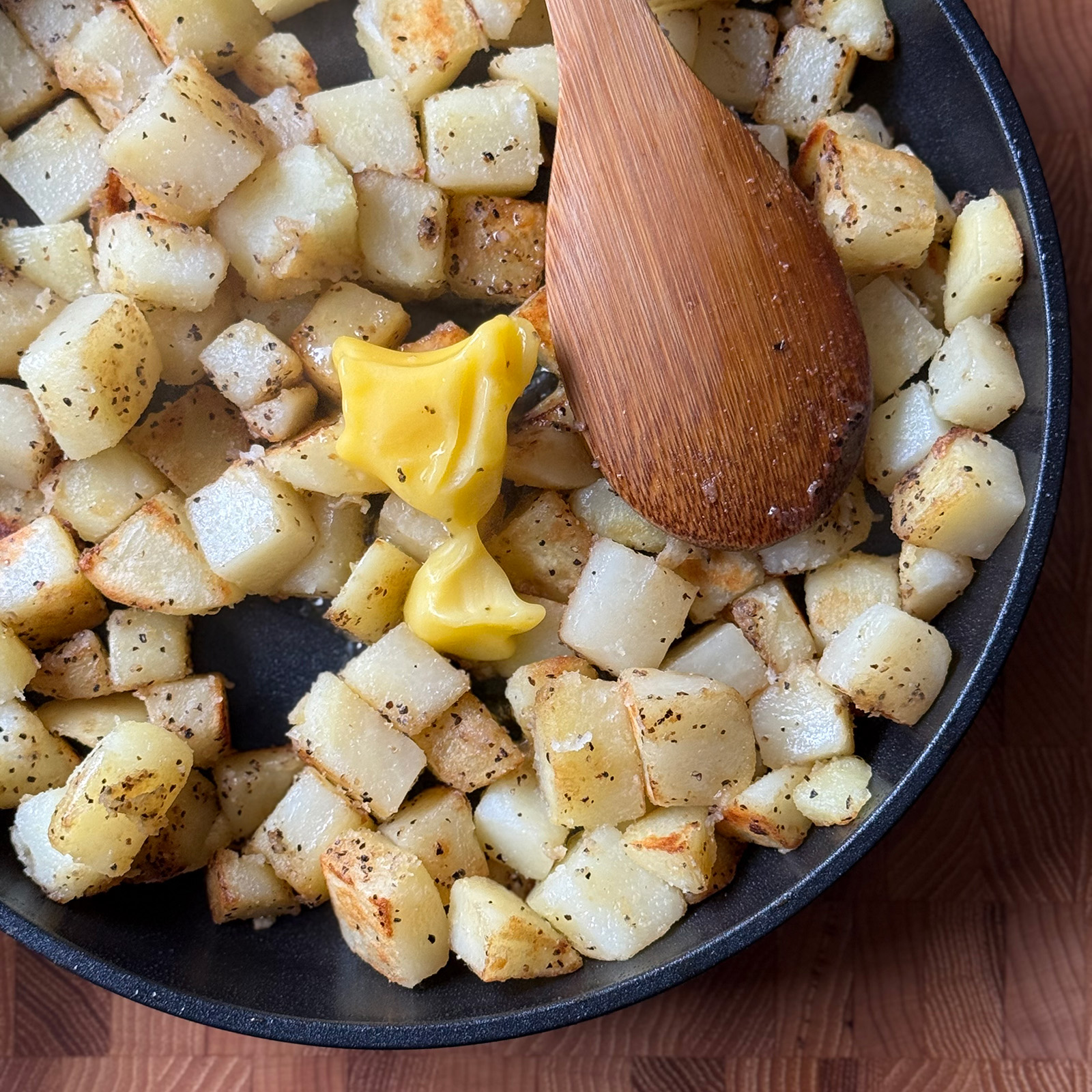 A spoonful of ghee melting into seasoned, browning potato cubes in a skillet with a wooden spoon.