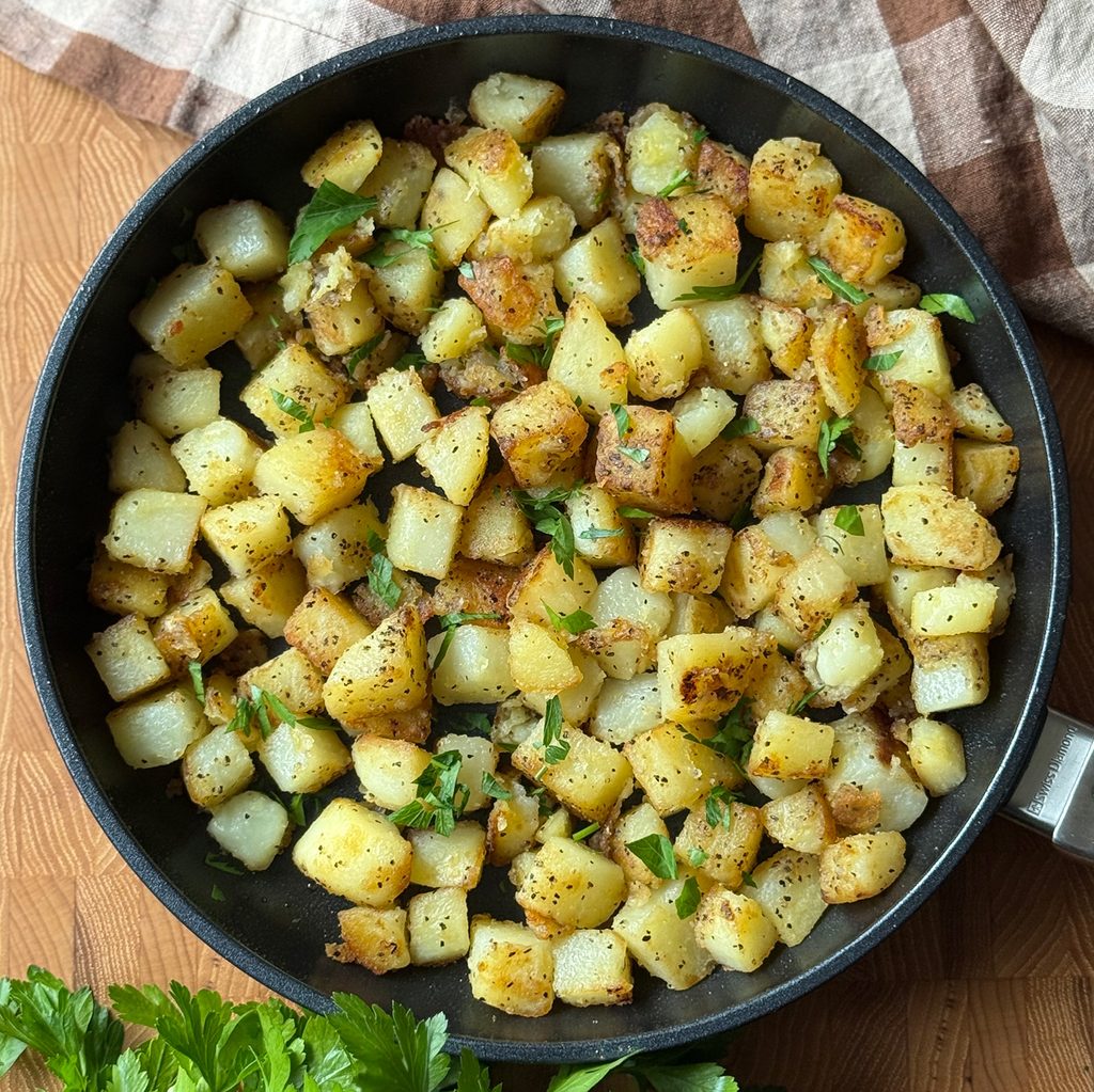 Home Fries in a skillet, garnished with fresh chopped parsley. Parsley and a checkered napkin are tucked at the edges of the pan.