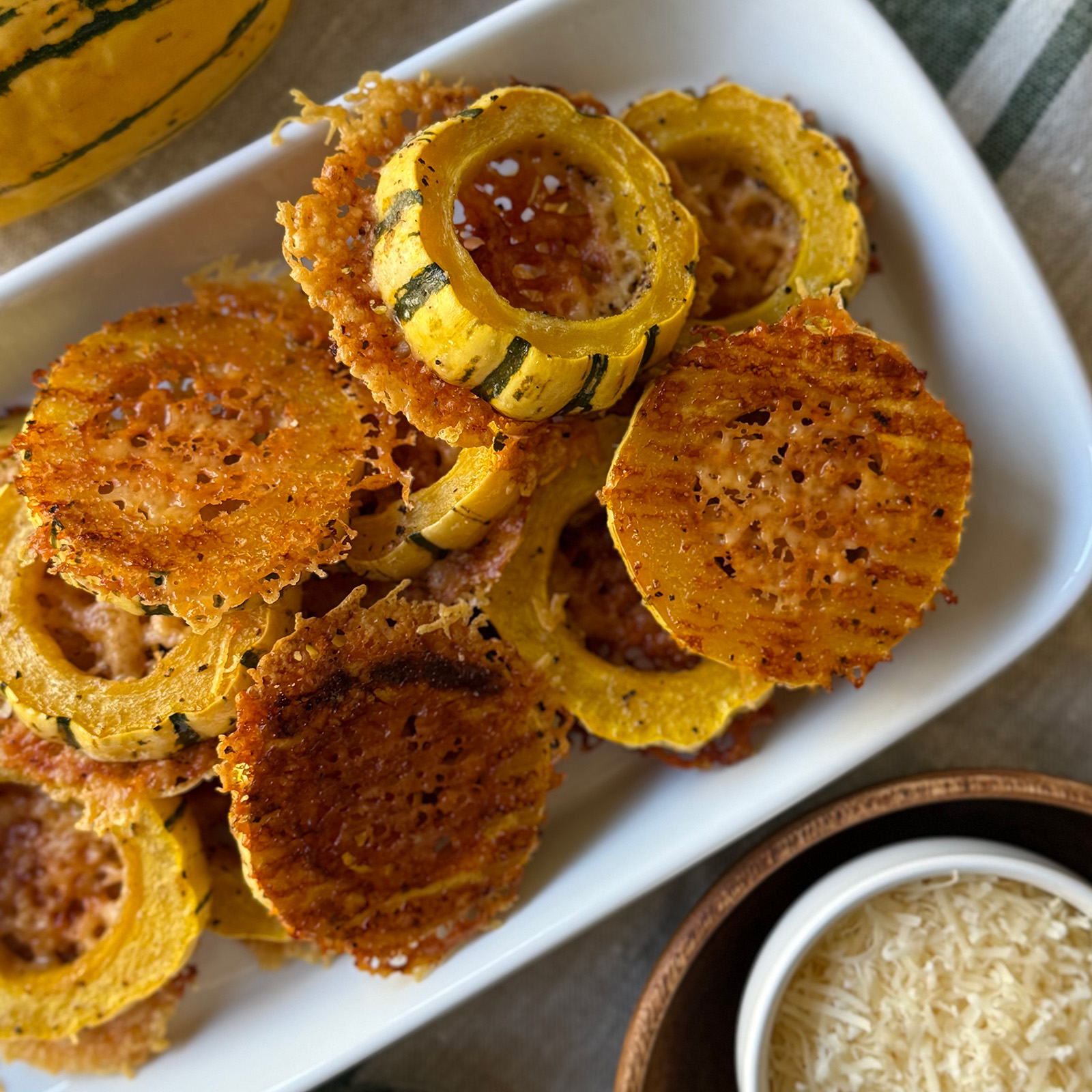 Crispy Parmesan delicata squash rings on a white platter, some flipped to show a lacy golden cheese crust, with a bowl of grated Parmesan on the side.