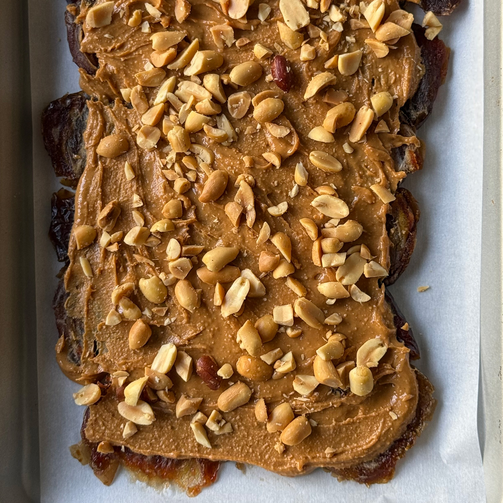 Peanut butter spread over pressed dates and topped with chopped roasted peanuts on a sheet pan.