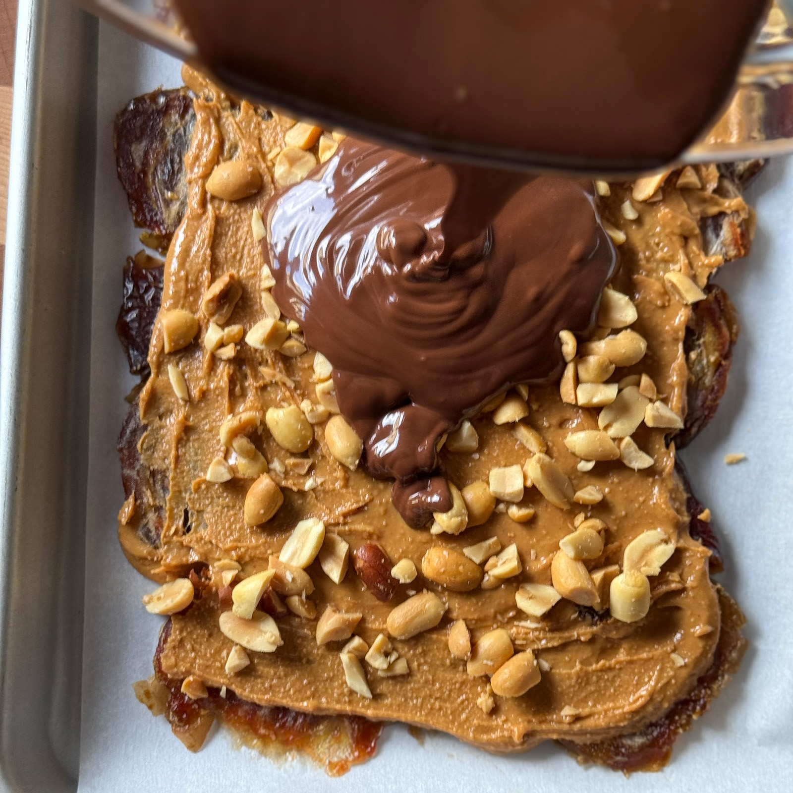Melted chocolate being poured over the peanut butter and chopped peanut layer.