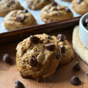 Two pumpkin chocolate chip cookies resting on a wooden board, with a baking sheet of more cookies in the background.