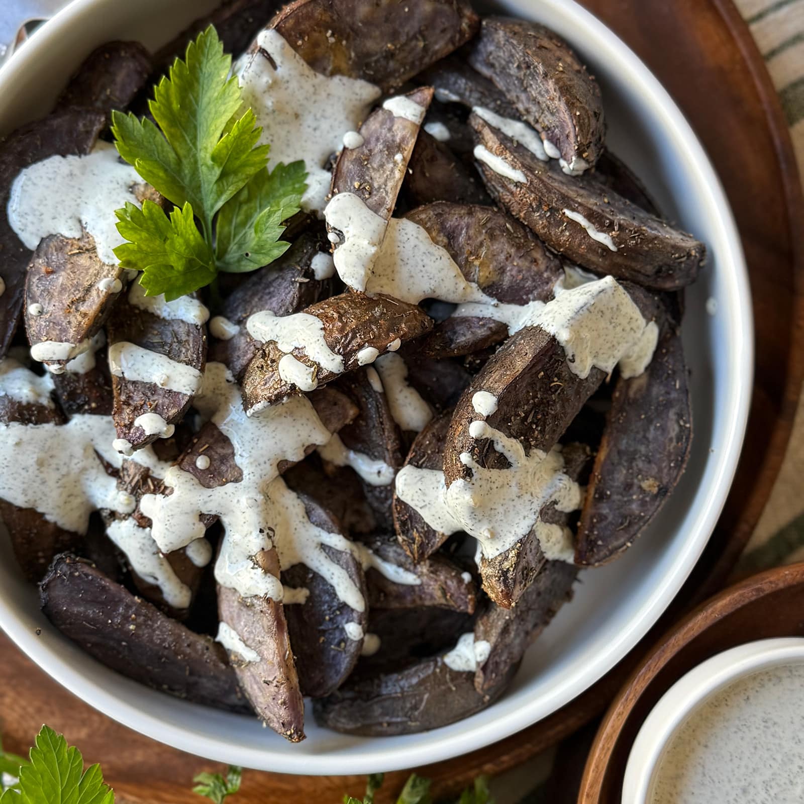 A white bowl filled with roasted purple potato wedges drizzled with creamy ranch dressing and garnished with a sprig of parsley.
