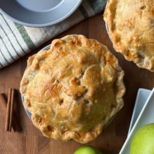 Two baked small apple pies with golden-brown crusts on a wooden surface, accompanied by cinnamon sticks and green apples.