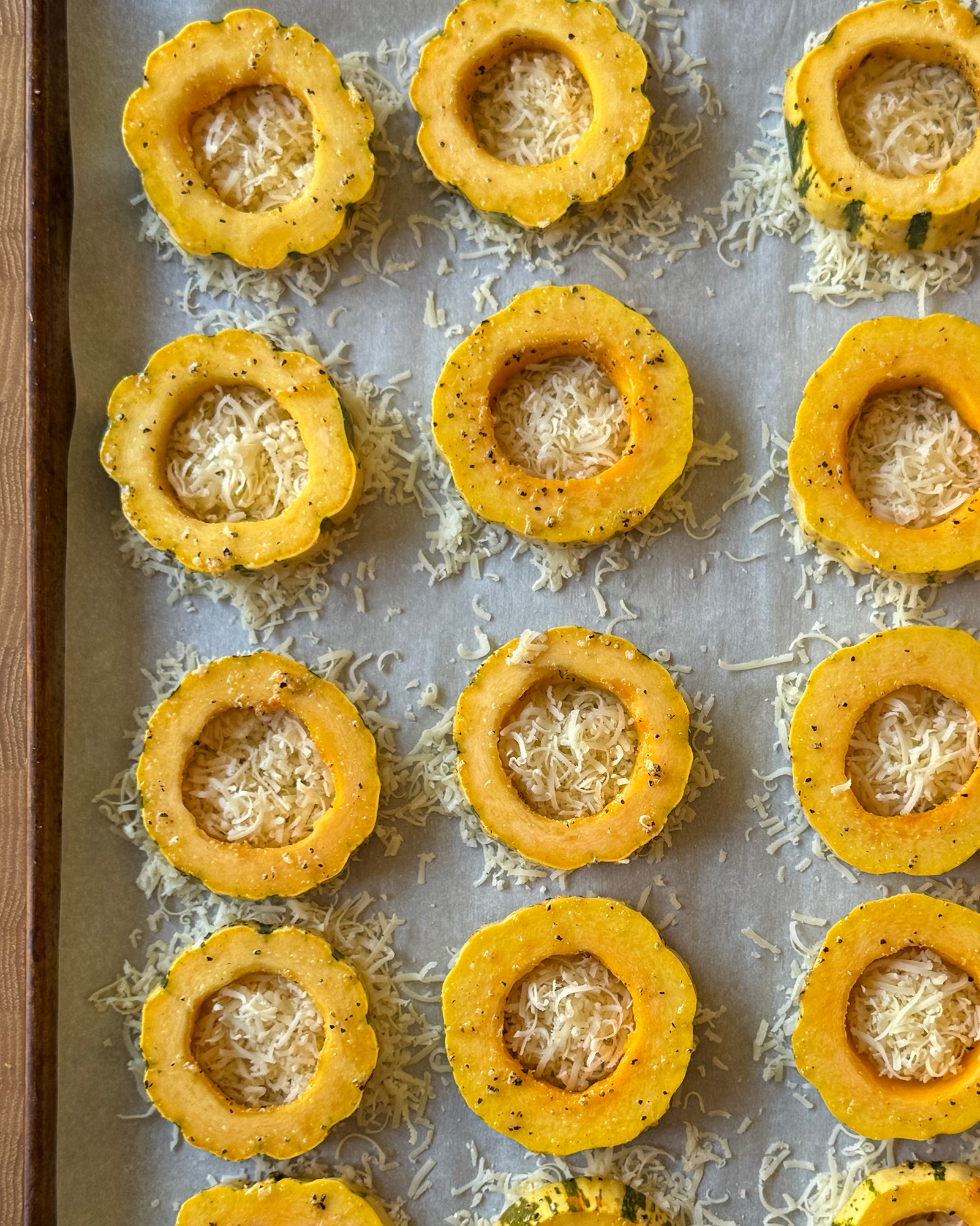 Seasoned delicata squash rings set on mounds of grated Parmesan on a parchment-lined baking sheet, ready for the oven.